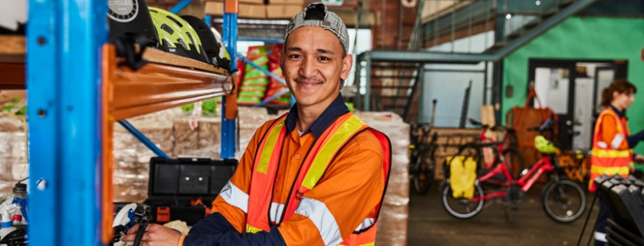 Young man in orange high-vis shirt in warehouse, wearing a backwards cap and smiling to camera