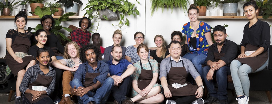 Group of young people in aprons on a large lounge smiling to camera with plants hanging behind them