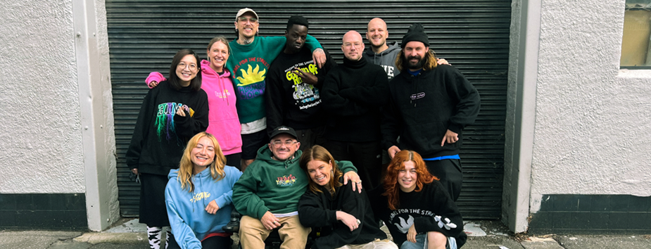 A diverse group of eleven young people smiling in front of a garage door on the street outside