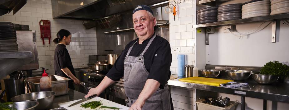 Male chef in kitchen standing in front of a cutting board with female chef in the background